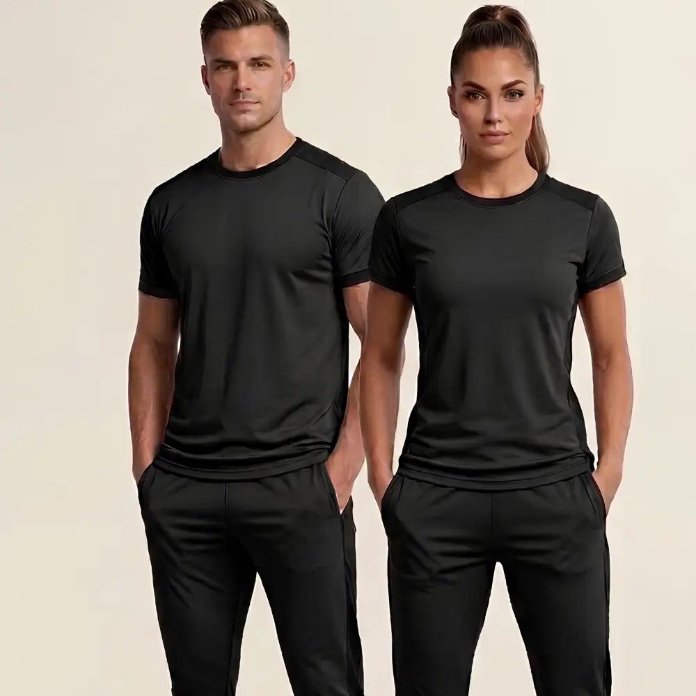 A man and a woman modeling matching black athletic short-sleeve crewneck shirts and black pants against a light beige studio background.
